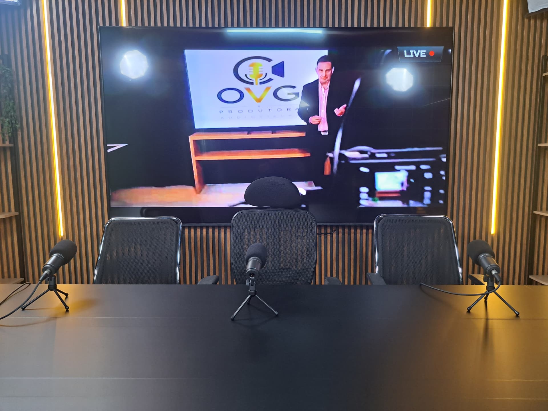 Interior do estúdio da OVG com set de podcast, microfones e telão em transmissão ao vivo