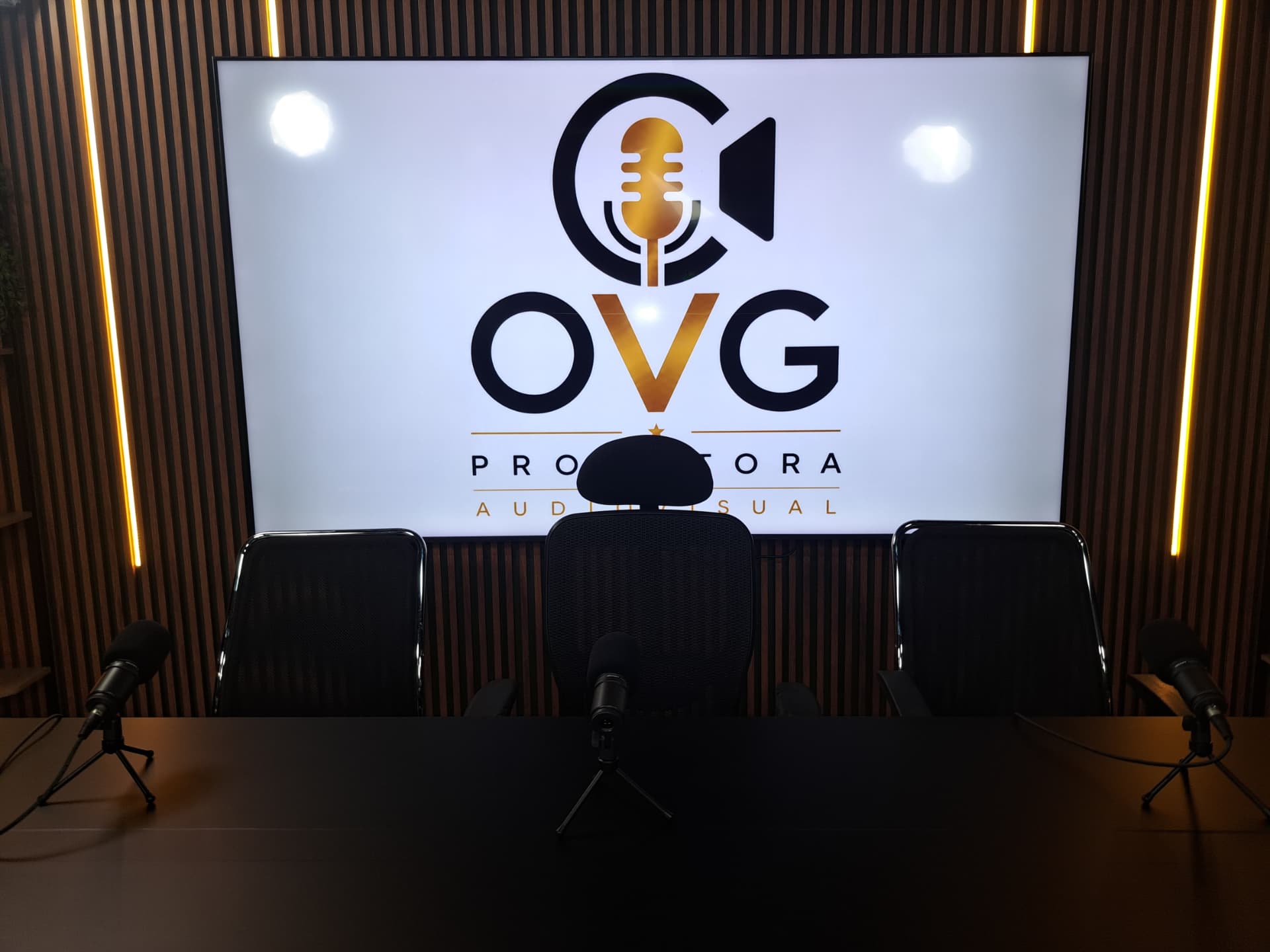 Sala de podcast da OVG com três microfones profissionais e telão com a marca OVG